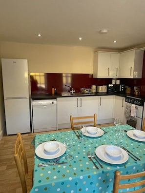 A compact kitchen with white cabinets, a refrigerator, a dishwasher, and a dining area with a polka dot tablecloth.