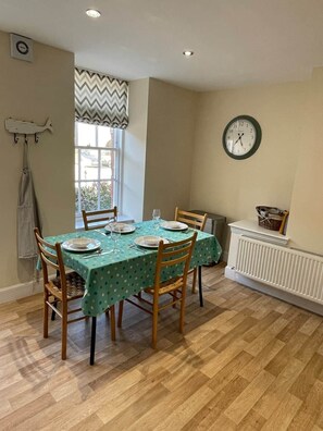 A dining area with a table set for four, wooden chairs, a clock on the wall, and a window with a chevron-patterned valance.