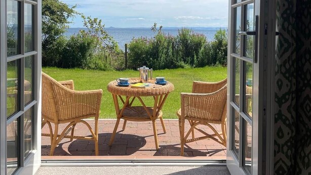 Glijdende glazen deuren geven uitkomst op een terras met een tafel en stoelen, een gazon en uitzicht op de zee.