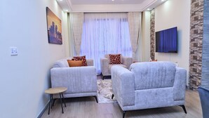 A living room with two sofas, a small table, a TV, and a window with curtains. - Tulivu By Edmor Suites (Nairobi)