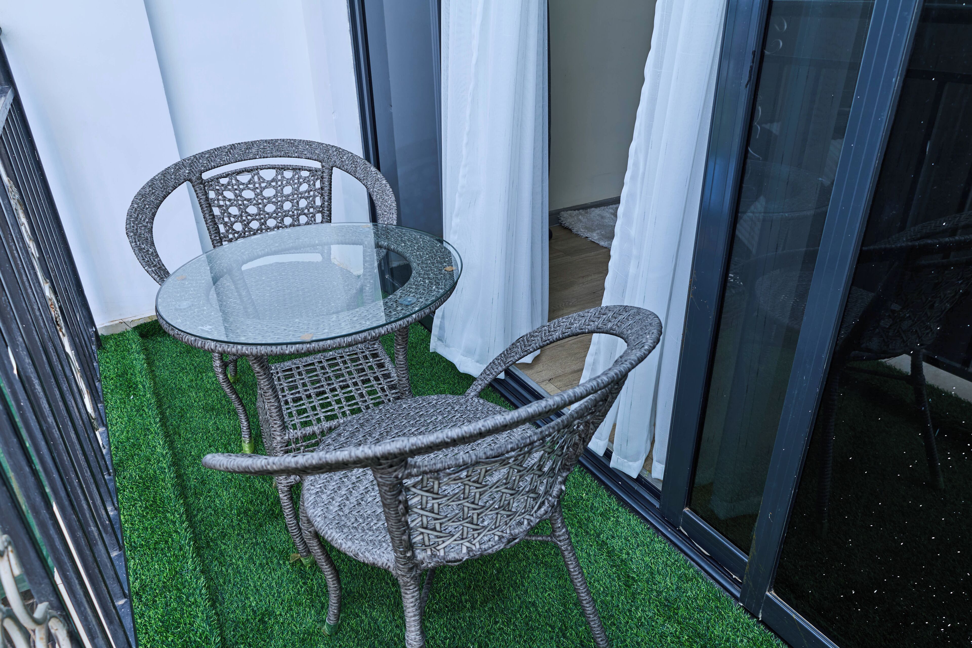 A balcony with a glass table and wicker chairs, surrounded by white curtains and a green artificial turf floor.