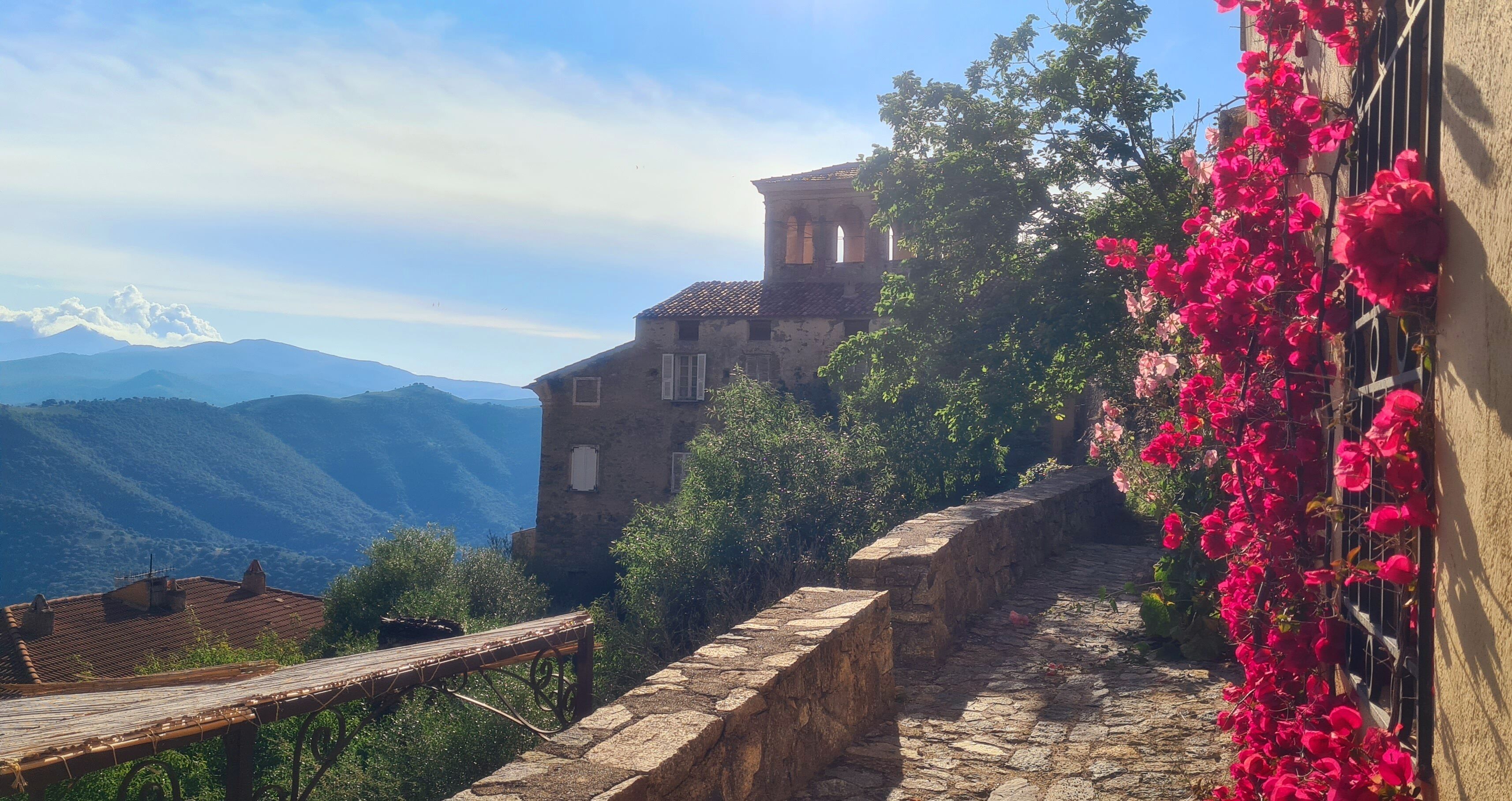 Un sentiero in pietra conduce a un edificio rustico con un campanile, circondato da vegetazione lussureggiante e montagne sullo sfondo.