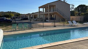 Vista de la piscina con un edificio de dos pisos y un balcón, zona vallada con sillones de descanso y un cielo azul despejado.