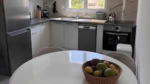 Cocina moderna con refrigerador de acero inoxidable, lavavajillas, estufa y pileta. Una ventana proporciona luz natural y vistas al exterior. En primer plano, una mesa con una cesta de frutas.