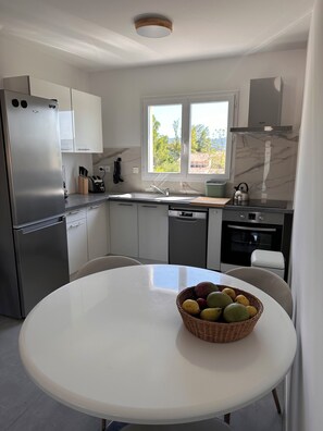 A modern kitchen with a stainless steel refrigerator, a dishwasher, a stove, and a sink. There is a window providing natural light and a view of the outdoors. A table with a basket of fruits is in the foreground. - Ô Gorges-a 5-minute walk from the river and the Ardèche gorges (Vallon-Pont-d'Arc)