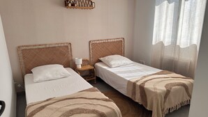 Habitación doble con dos camas individuales, una mesita de noche con lámpara, cabecero tejido y vistas al exterior a través de cortinas translúcidas.