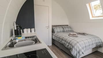 Habitación compacta con kitchenette, una cama con colcha de cuadros y ventana con vista a árboles.
