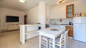 Een moderne keuken met witte kasten, een eethoek met een witte tafel en stoelen, een wandgemonteerde flatscreen televisie en een ingebouwde magnetron.