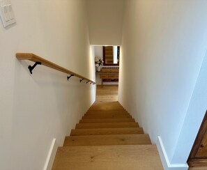 Una escalera de madera con barandilla, que lleva a una habitación con piso de madera y pared blanca.