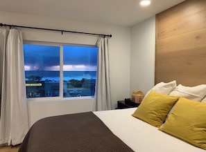 A bedroom with a large bed, a window with curtains, a nightstand, and a view of the outside. - Cloud Ninth - Ocean View Yachats (Yachats)