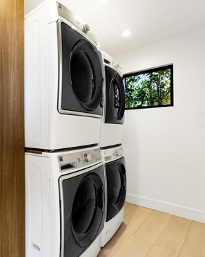 A laundry room with stacked washers and dryers, a wooden door, and a window with a view of trees. - Saint Ives Reserve - Sunset Strip Retreat w/ Epic City-to-Ocean Views (Los Angeles)