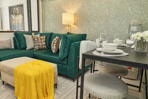 Un sofá verde con cojines estampados, una manta amarilla, una mesa de comedor con platos y vasos, y una lámpara fijada a la pared.