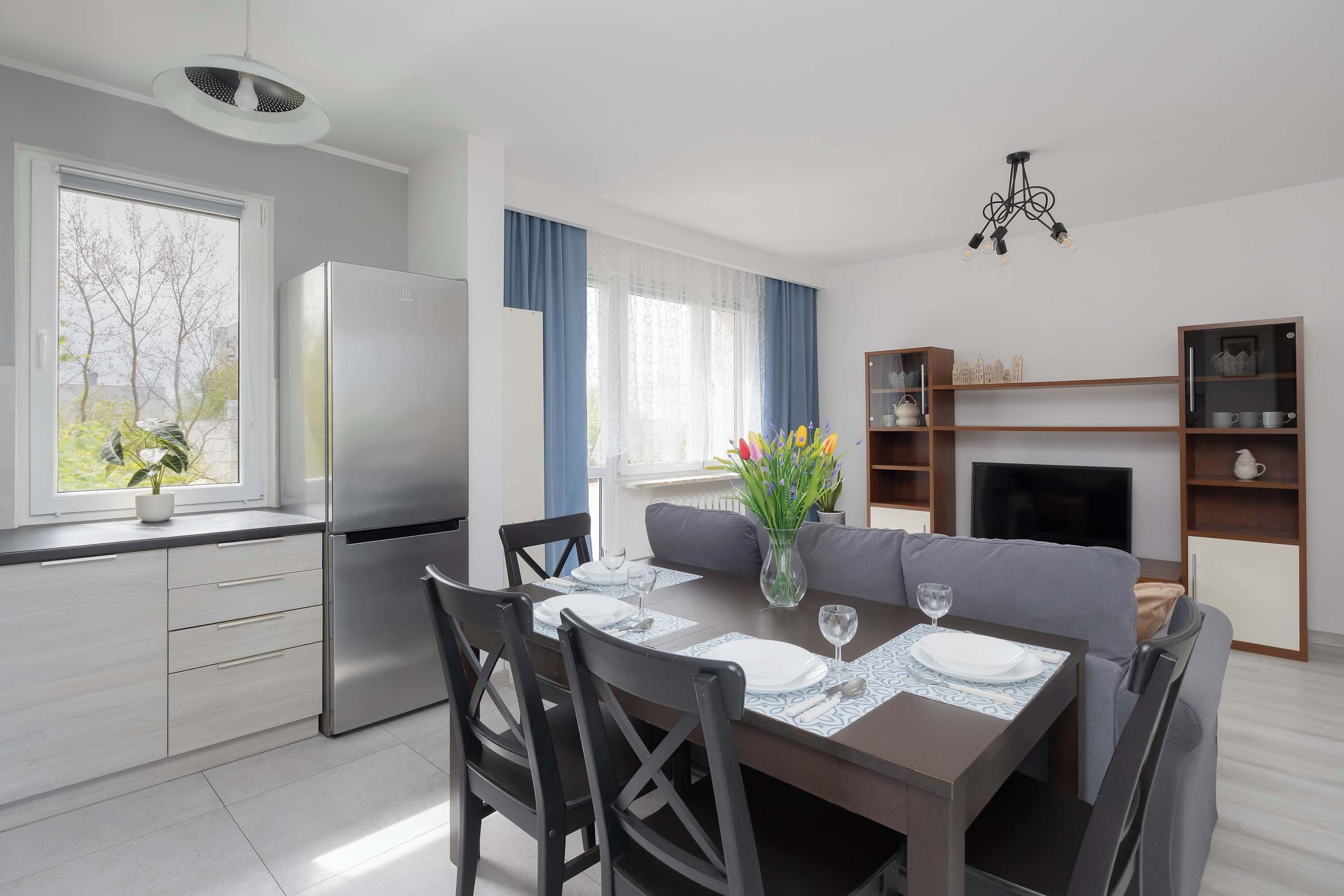 A modern kitchen with a refrigerator, dining table set for two, and a sofa in the living area.