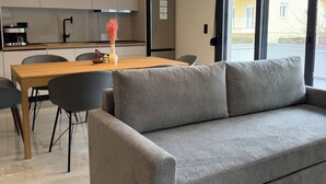 Uma sala de estar moderna com um sofá cinzento, uma mesa de jantar de madeira e uma área de cozinha com frigorífico e máquina de café.