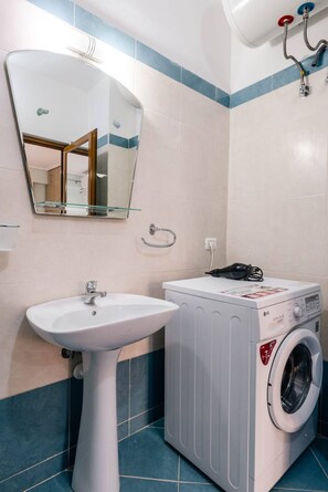 A bathroom with a pedestal sink, a washing machine, a mirror, and a towel rack. - Albania Dream Saranda Apartments (Sarandë)