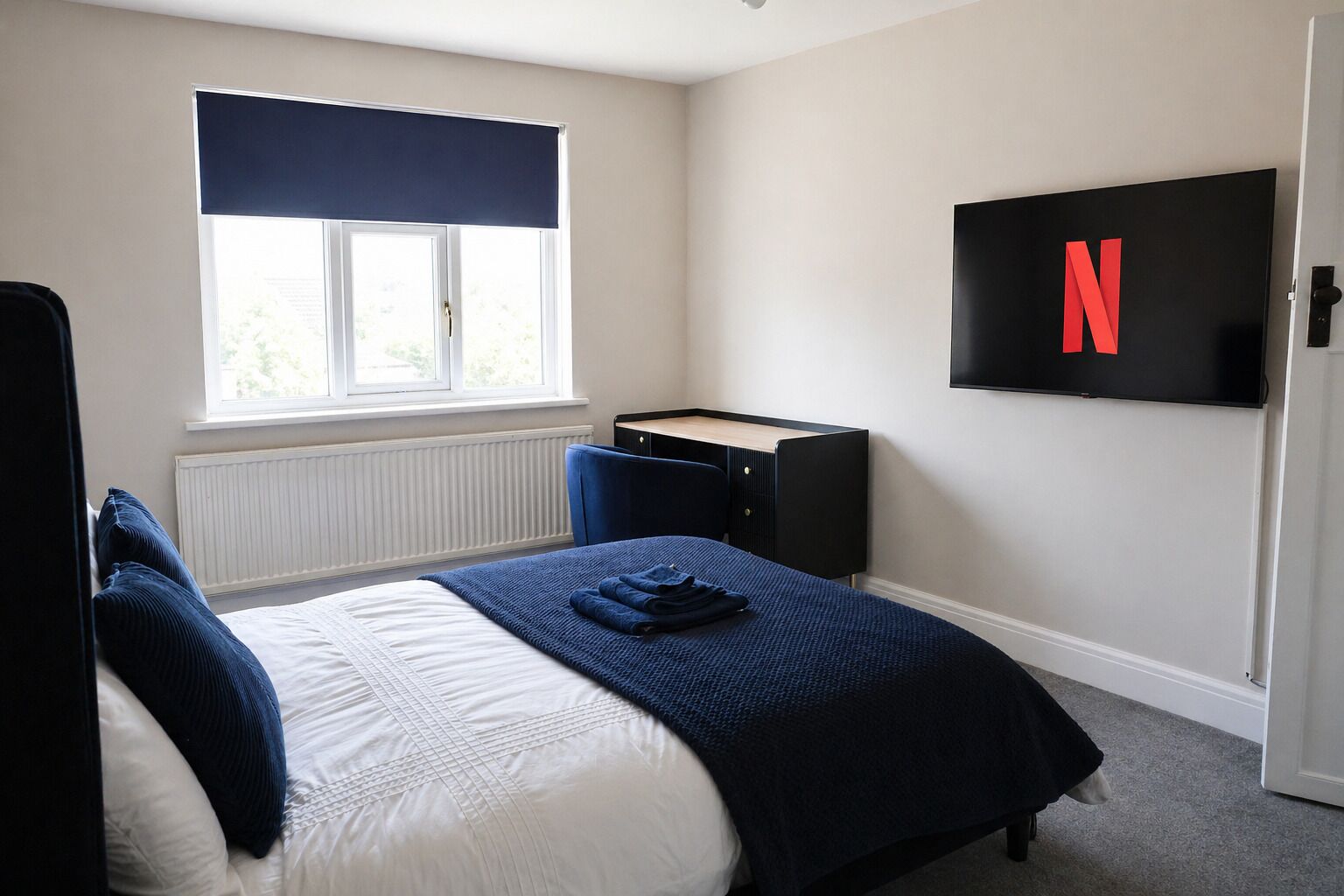 Un dormitorio con cama, escritorio, silla, radiador, ventana con persiana y televisor con logotipo de Netflix.