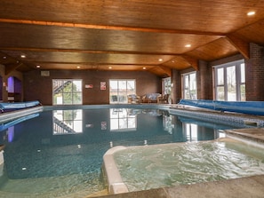 Una piscina interior con agua cristalina, vigas de techo de madera y una zona de estar con sillas y sofá.