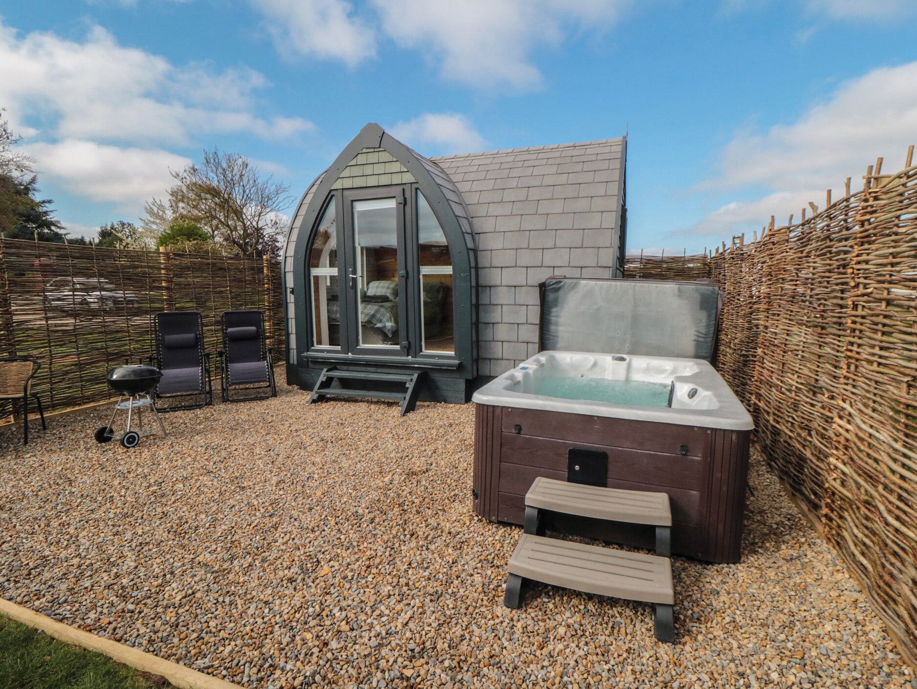 A cozy outdoor area with a hot tub, two lounge chairs, and a small step leading to a glass-enclosed room.