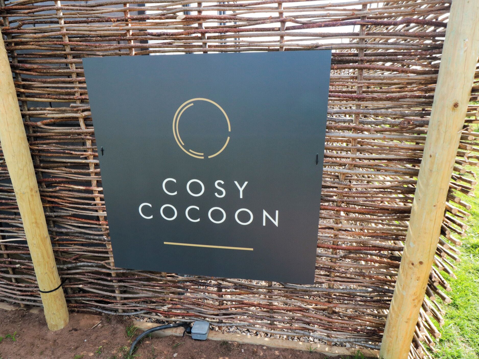 Un letrero con el texto 'COSY COCOON' sobre un fondo oscuro, sujeto a una estructura de madera.