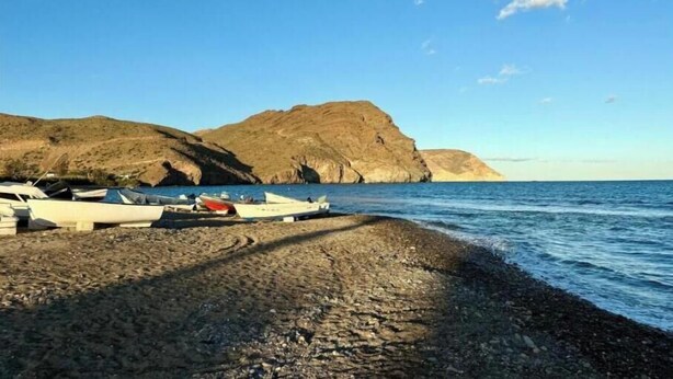 Una playa rocosa con barcos amarrados, cielo despejado y montañas al fondo.