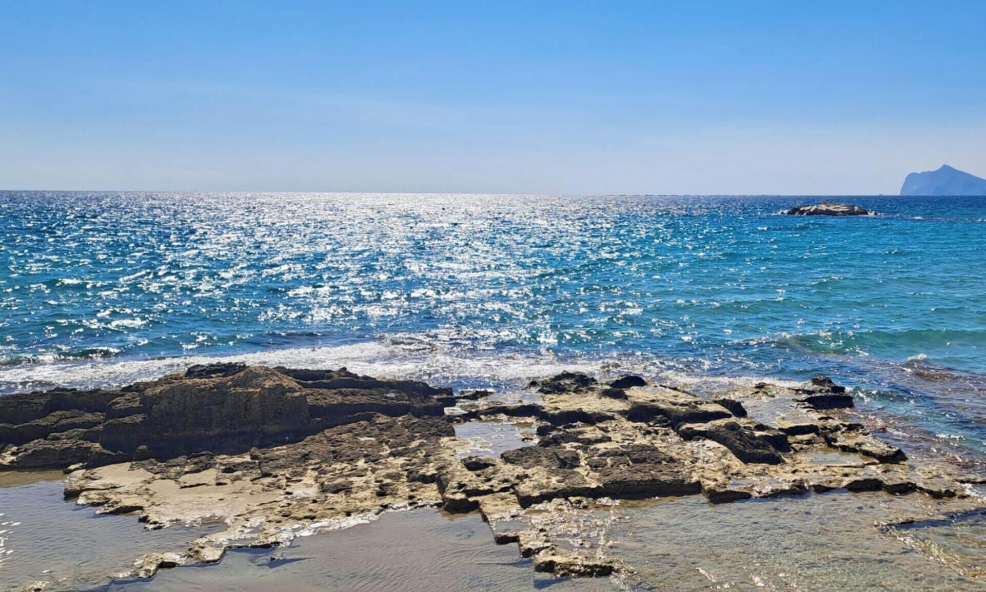A coastal scene with clear blue water, rocky shoreline, and a distant mountain.