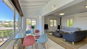 A modern living room with a glass dining table, colorful chairs, a grey sofa, and a view of the city. - Elegant Villa in Moraira for 6 · Private Pool Close to Town – ALMARE (Teulada)