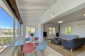 A modern living room with a glass dining table, colorful chairs, a grey sofa, and a view of the city. - Elegant Villa in Moraira for 6 · Private Pool Close to Town – ALMARE (Teulada)
