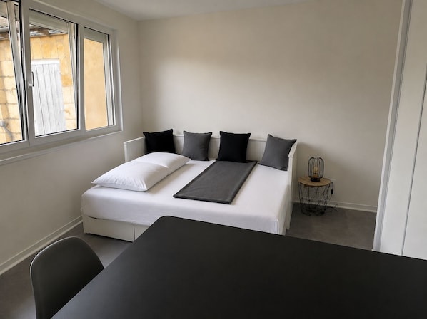 Ein Einzelbett mit weißer Matratze und schwarzen Kissen, ein Nachttisch mit Lampe sowie ein Fenster mit Blick auf ein Gebäude.