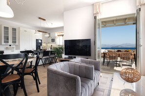 A modern living room with a flat-screen TV, grey sofa, dining area, kitchen, and a balcony with a view of the sea. - Sweet View (Paros)