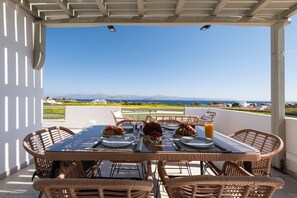 A covered outdoor dining area with wicker furniture, a table set with plates, glasses, and a fruit bowl, overlooking a scenic landscape. - Sweet View (Paros)