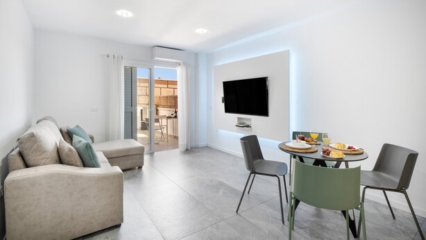 Ein modernes Wohnzimmer mit Sofa, Esstisch, Stühlen, einem Flachbildfernseher und einer durch eine offene Tür einsehbaren Küche.