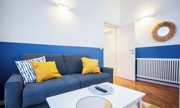 Een kamer met een grijze bank, twee gele kussens, een witte tafel met een afstandsbediening en een witte radiator.