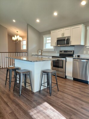 A modern kitchen with white cabinets, stainless steel appliances, and a central island with stools. - Start your Summer with a Vaca at the Beach Garden Bungalow (Poynette)