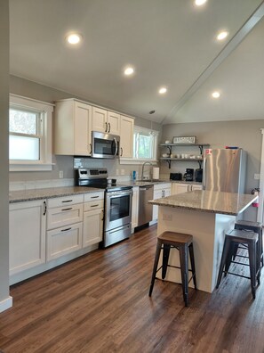 A modern kitchen with white cabinets, stainless steel appliances, a wooden floor, and a bar area with stools. - Start your Summer with a Vaca at the Beach Garden Bungalow (Poynette)
