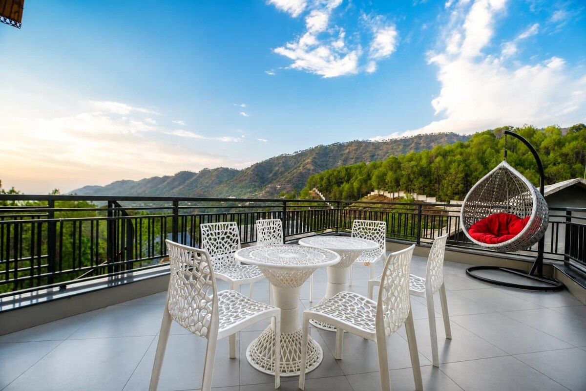 Eine Terrasse mit weißen Korbmöbeln, einem Hängesessel und Blick auf grüne Hügel.
