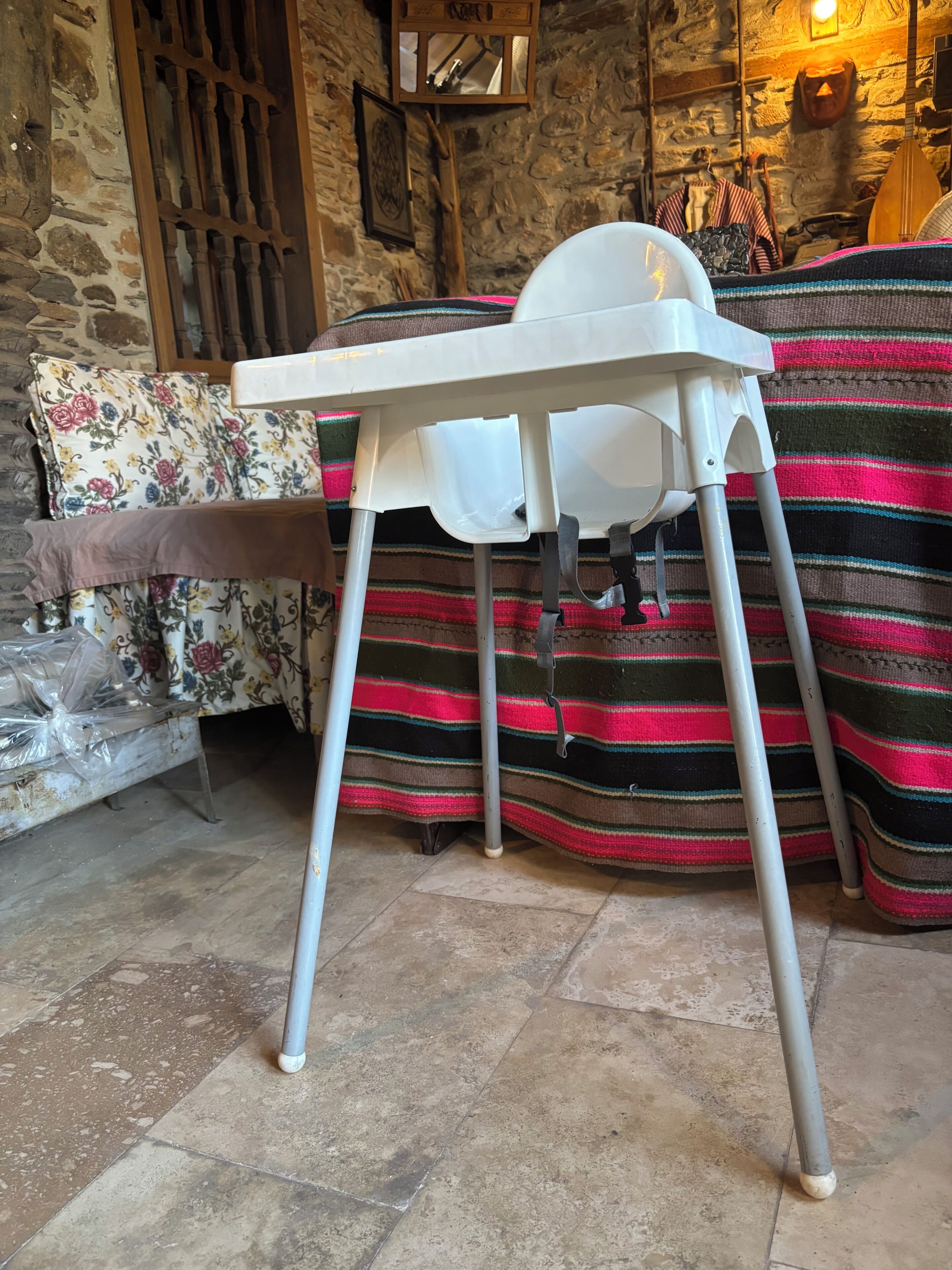 A white high chair with a tray table in a room with stone walls, a striped blanket, and a wooden door.
