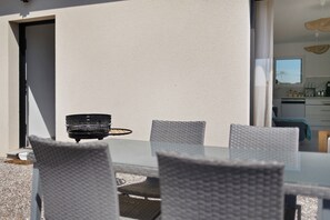 An outdoor dining area with a white table and wicker chairs, a black grill, and a kitchen visible through a glass door. - Holiday Home 'Maison Sci Entre Foret Et Mer' with Wi-Fi (Millebosc)