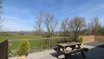 A patio with a picnic table and a small tree planter, overlooking a green field and trees.