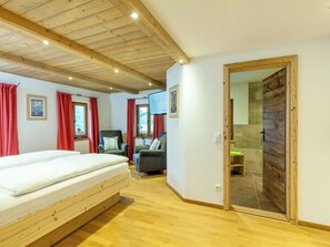 Ein Zimmer mit Holzboden, einem Bett, zwei Sesseln, einem Fernseher und einem durch eine Glastür einsehbaren Badezimmer.
