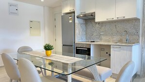 A modern kitchen with a glass dining table, white chairs, a built-in oven, and a stainless steel refrigerator.