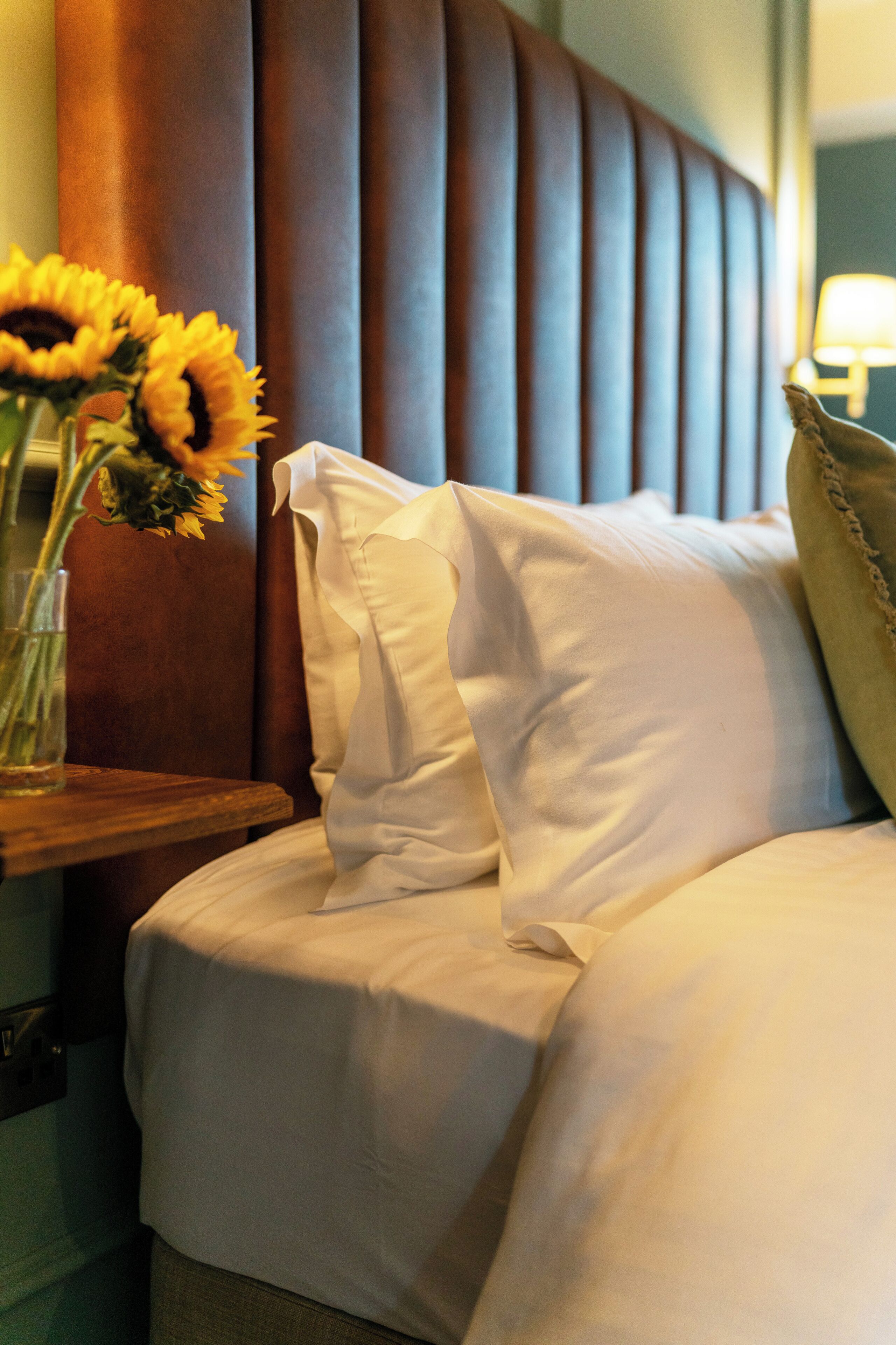 Una habitación de hotel con una cama, almohadas, un jarrón con girasoles y un cabecero de madera.