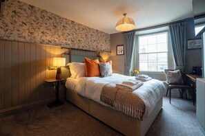 A hotel room with a large bed, a bedside table with a lamp, a chair, a window with curtains, and a wall with a floral pattern.