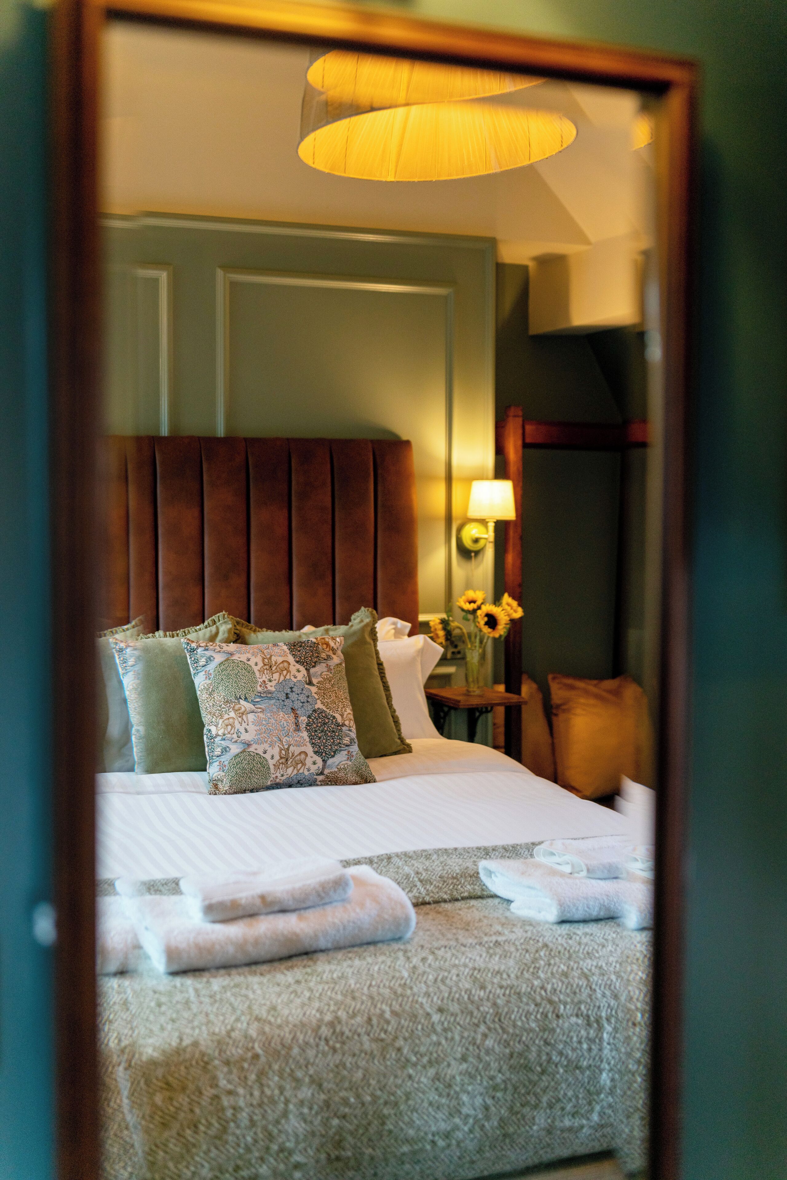 Una habitación de hotel con una cama, almohadas, una mesita de noche, una lámpara y un espejo que refleja la habitación.