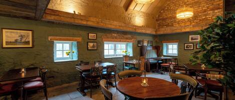 A rustic dining area with wooden tables and chairs, stone walls, and a large plant.