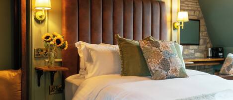A hotel room with a large bed, a headboard, wall-mounted lamps, a vase with sunflowers, and a stone wall.