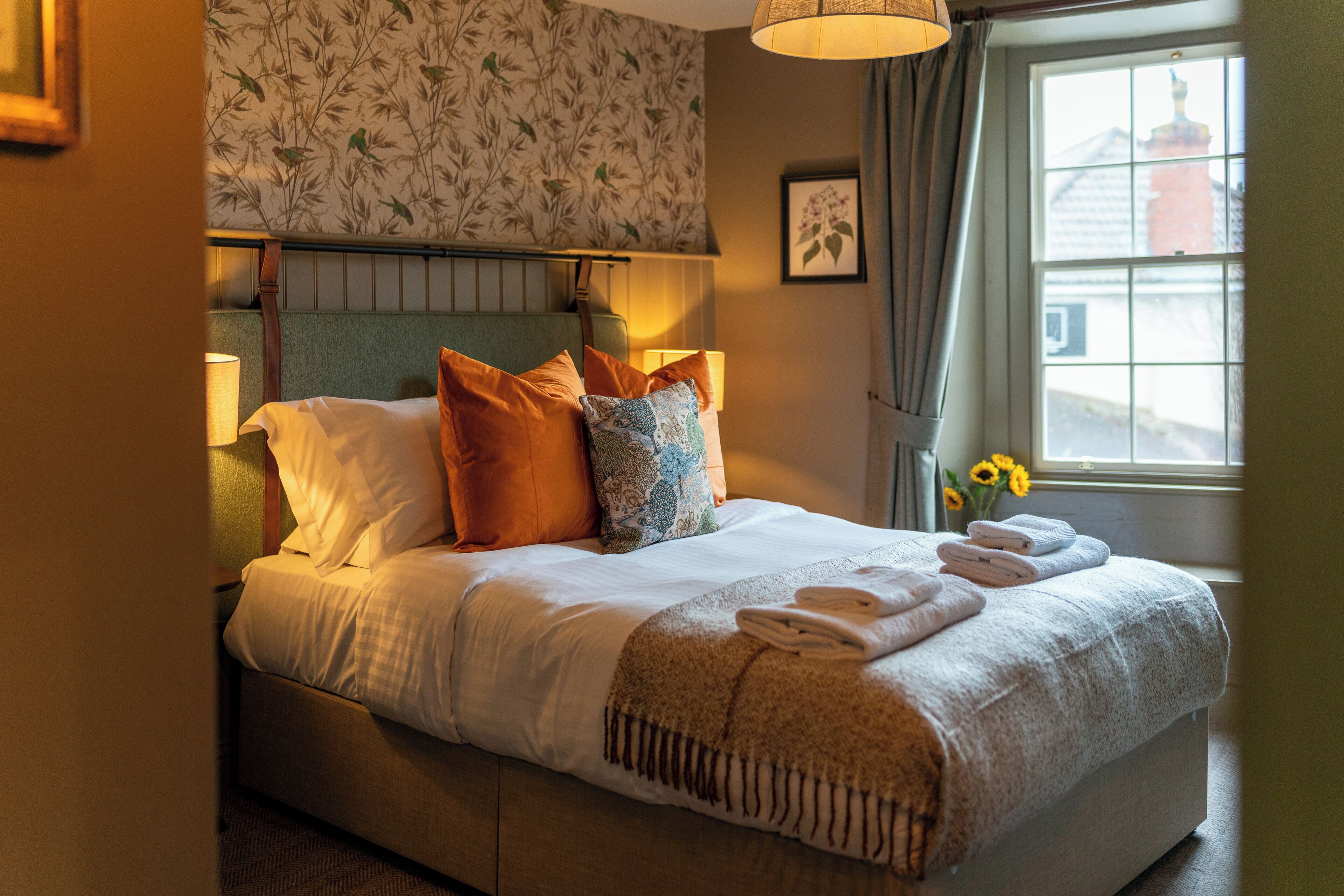 A neatly made bed with pillows, a bedside lamp, and a window with flowers.