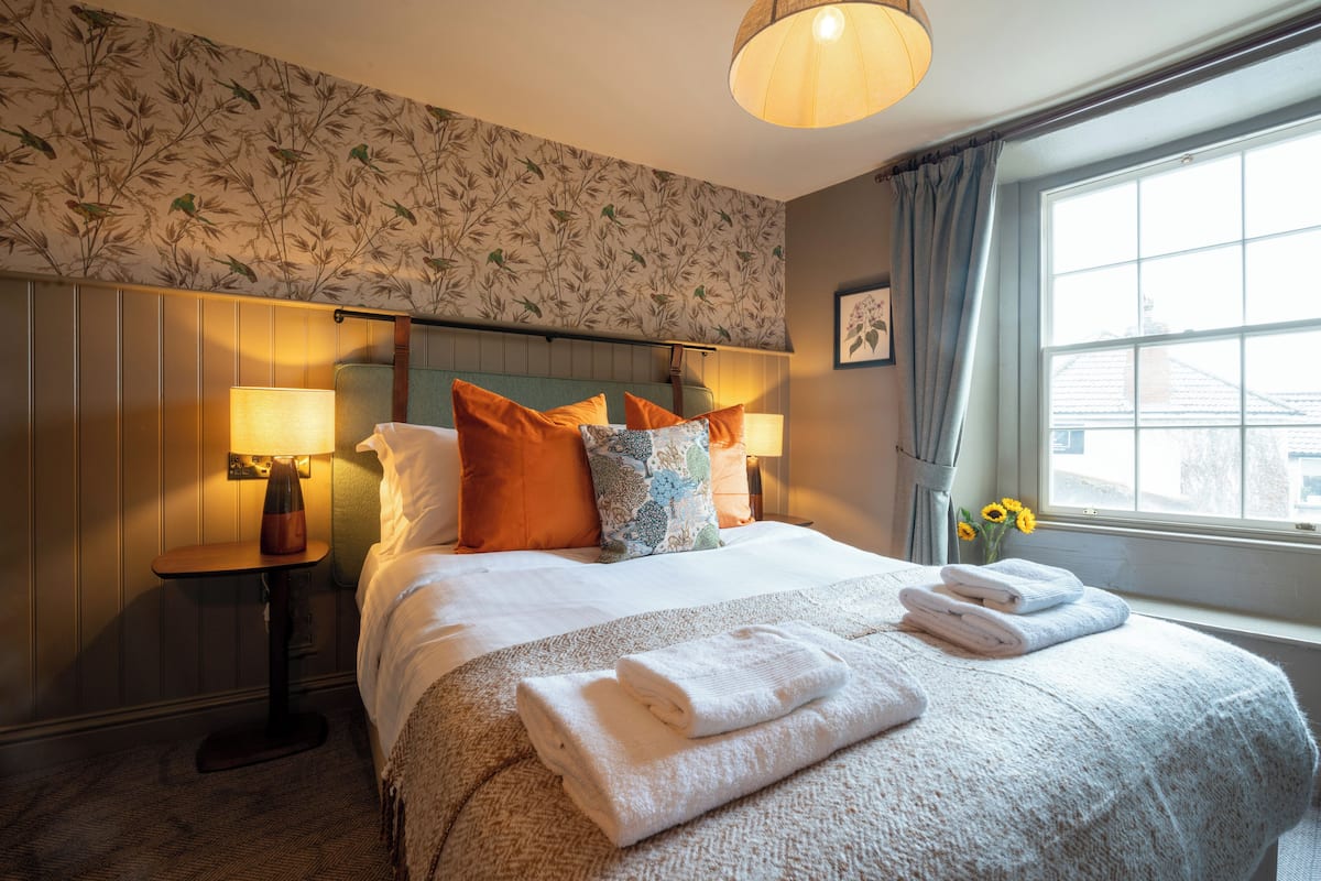 Habitación de hotel con una cama bien tendida, dos lámparas de noche, una ventana con cortinas y un estampado de flores en el papel tapiz.