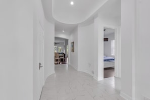A hallway with white walls and marble flooring leading to a bedroom with a bed and a window.