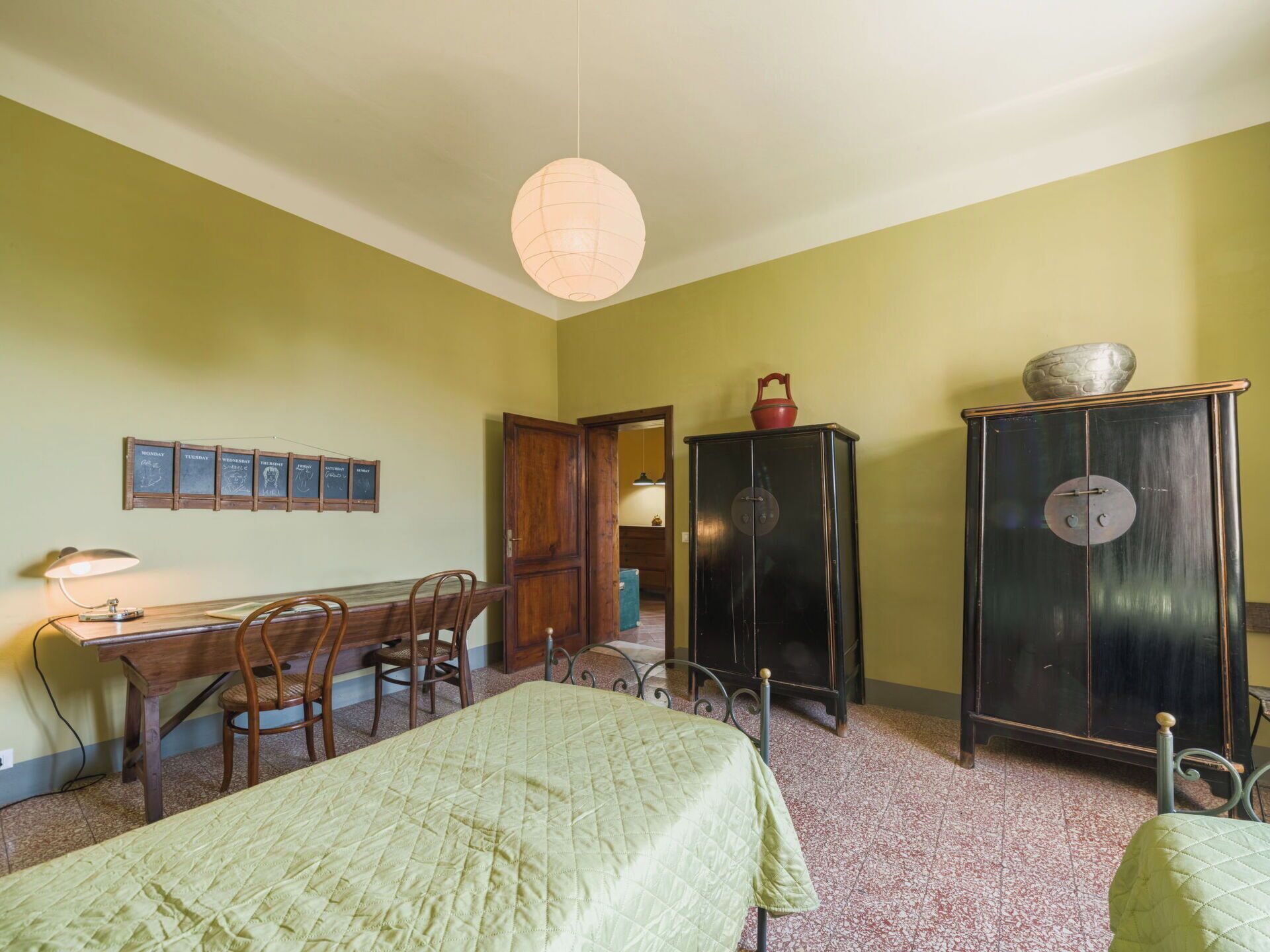 Une pièce comprenant un bureau, deux chaises, un lit avec une couette verte, une armoire et un luminaire fixé au mur.
