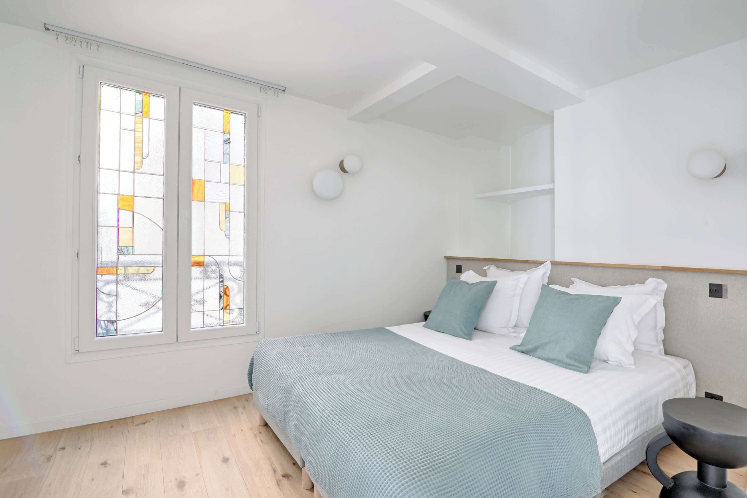 Een slaapkamer met een bed, een stoel, een raam met glas-in-lood en een plafond met zichtbare balken.